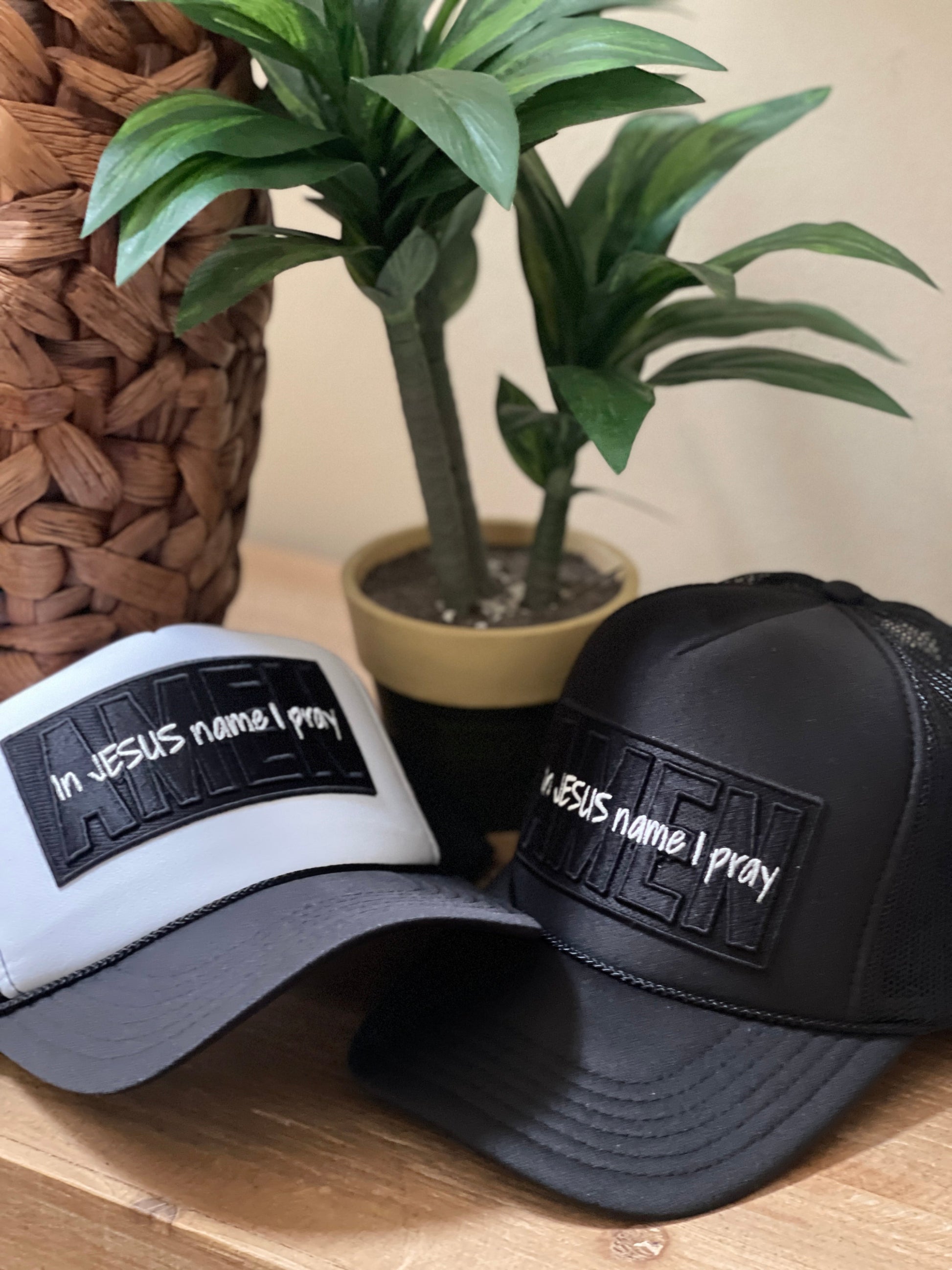 Two foam trucker hats, one white and one black, with an embroidered patch that reads 'In Jesus Name I Pray Amen,' placed on a wooden surface with a plant in the background.