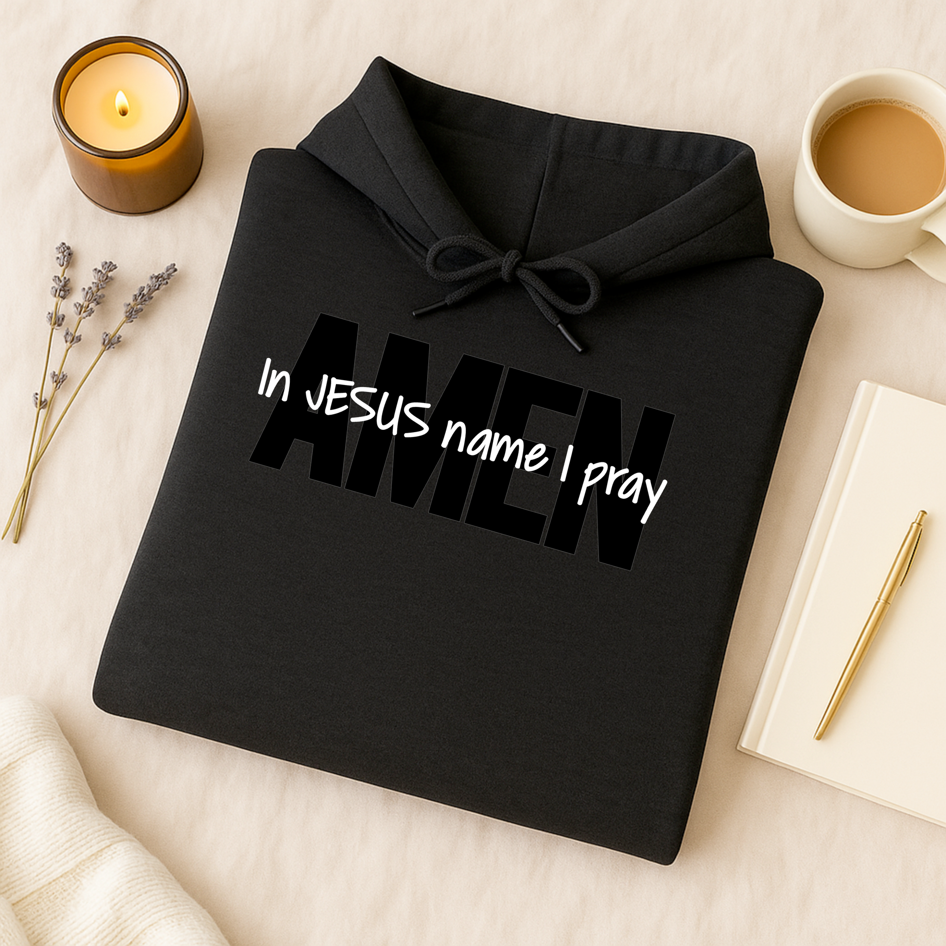 Folded black hoodie with the phrase “In Jesus Name I Pray” printed in white letters and “Amen” in bold black letters. The hoodie rests on a light background beside a warm cup of coffee, creating a cozy and faith-inspired flat lay aesthetic.